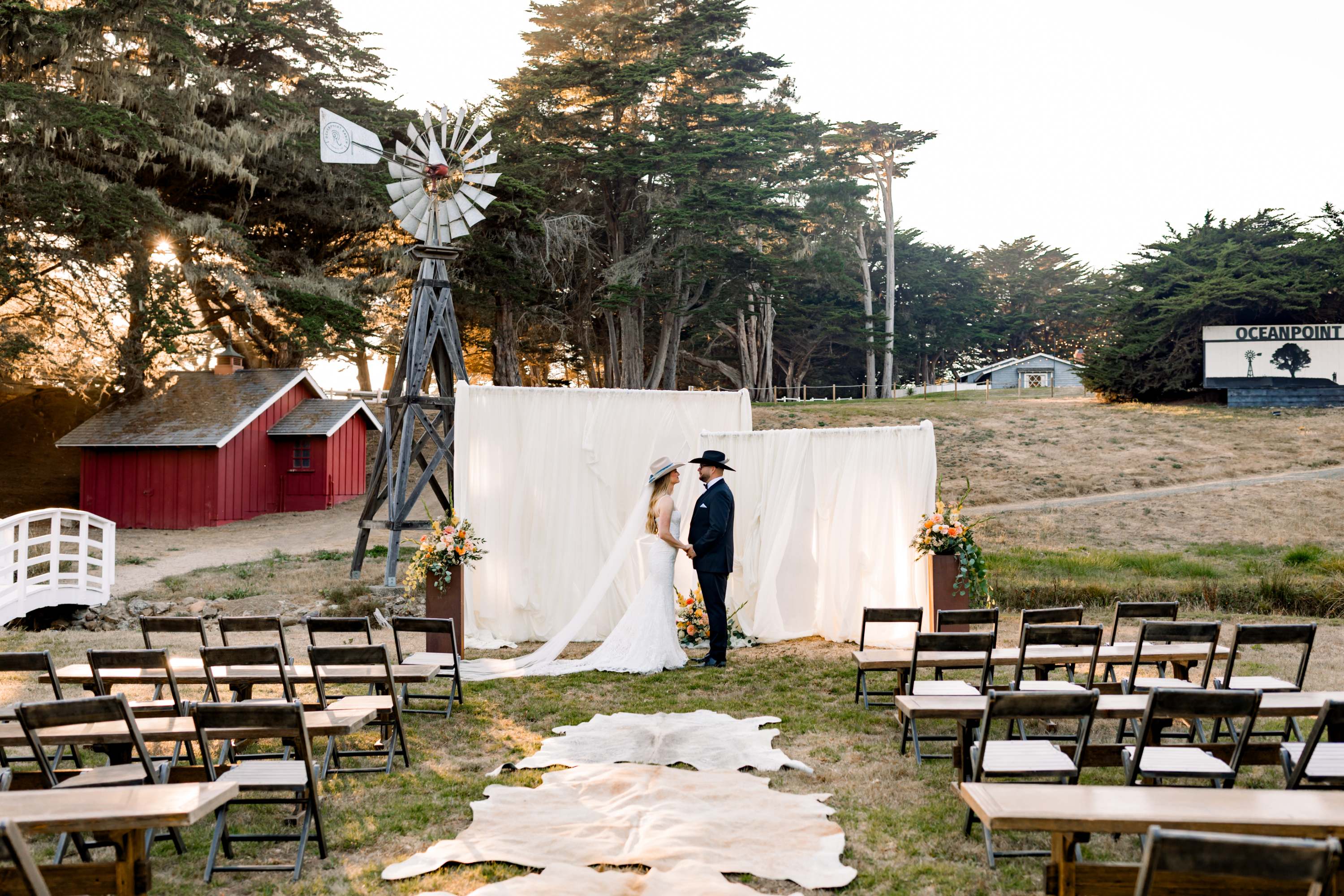 An outdoor wedding setup with a white backdrop, chairs, and a windmill in the background.