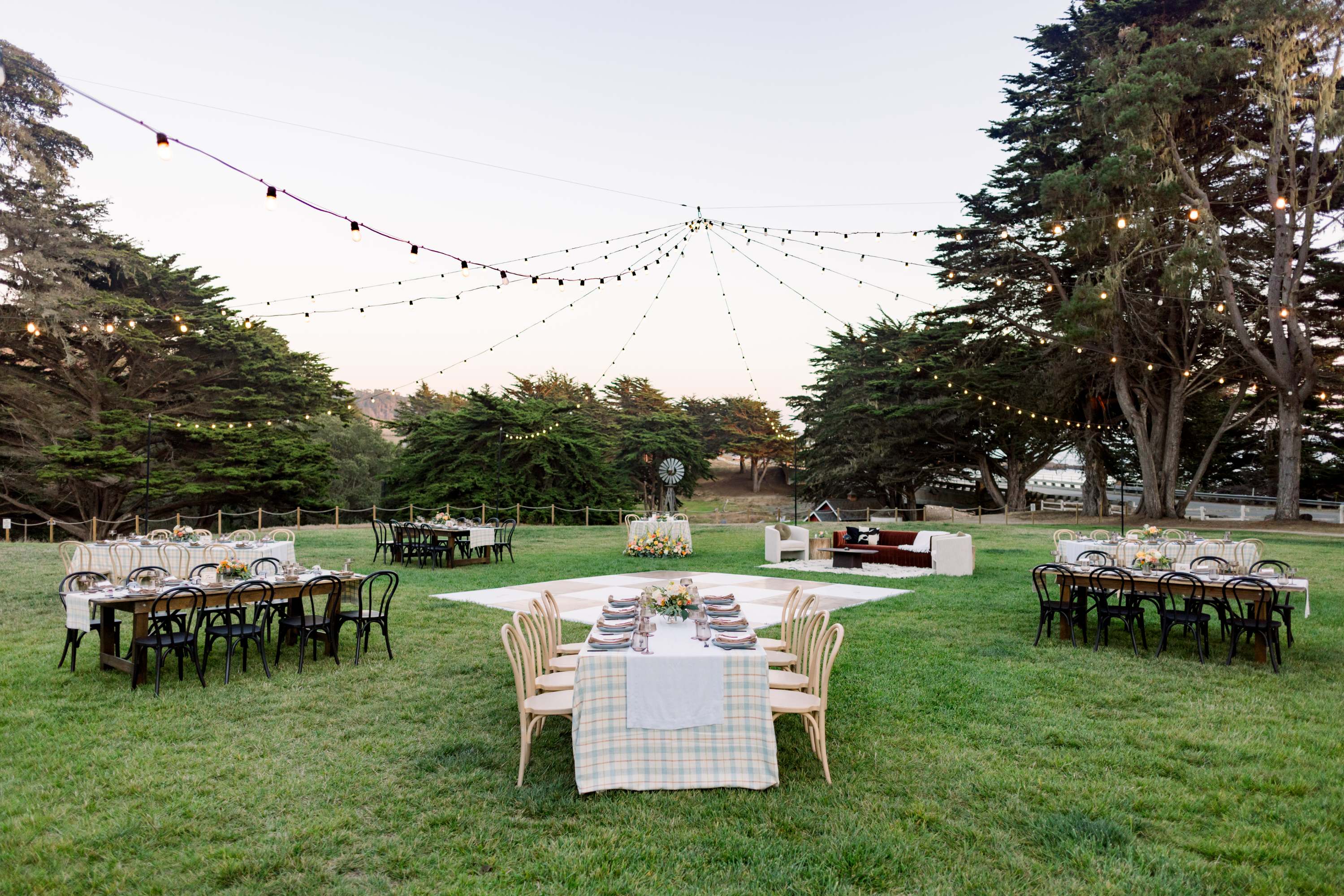 An outdoor event space with tables, chairs, and string lights set up on a grassy area surrounded by trees.