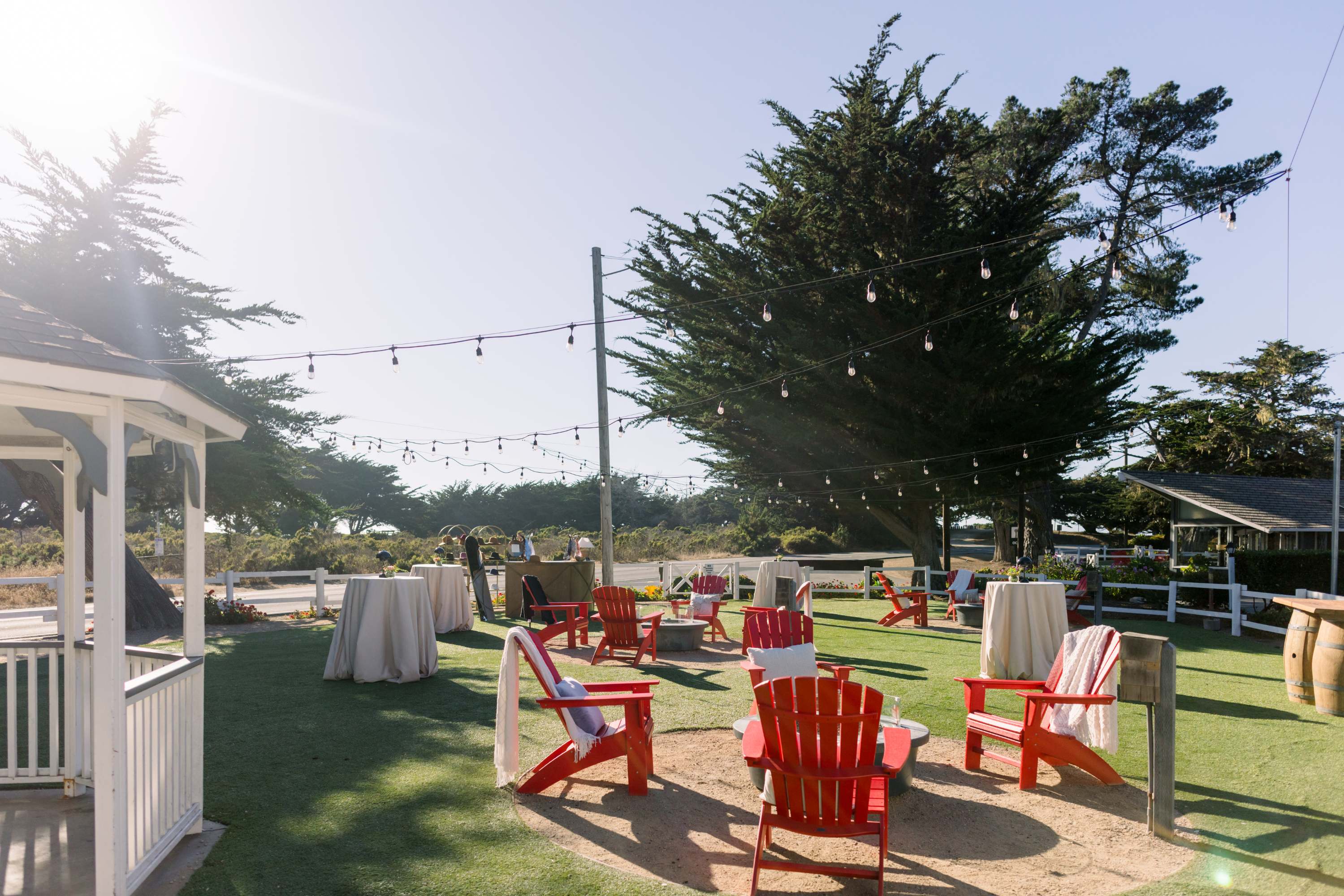 A well-manicured outdoor seating area with red Adirondack chairs, string lights, and tables set up for an event.