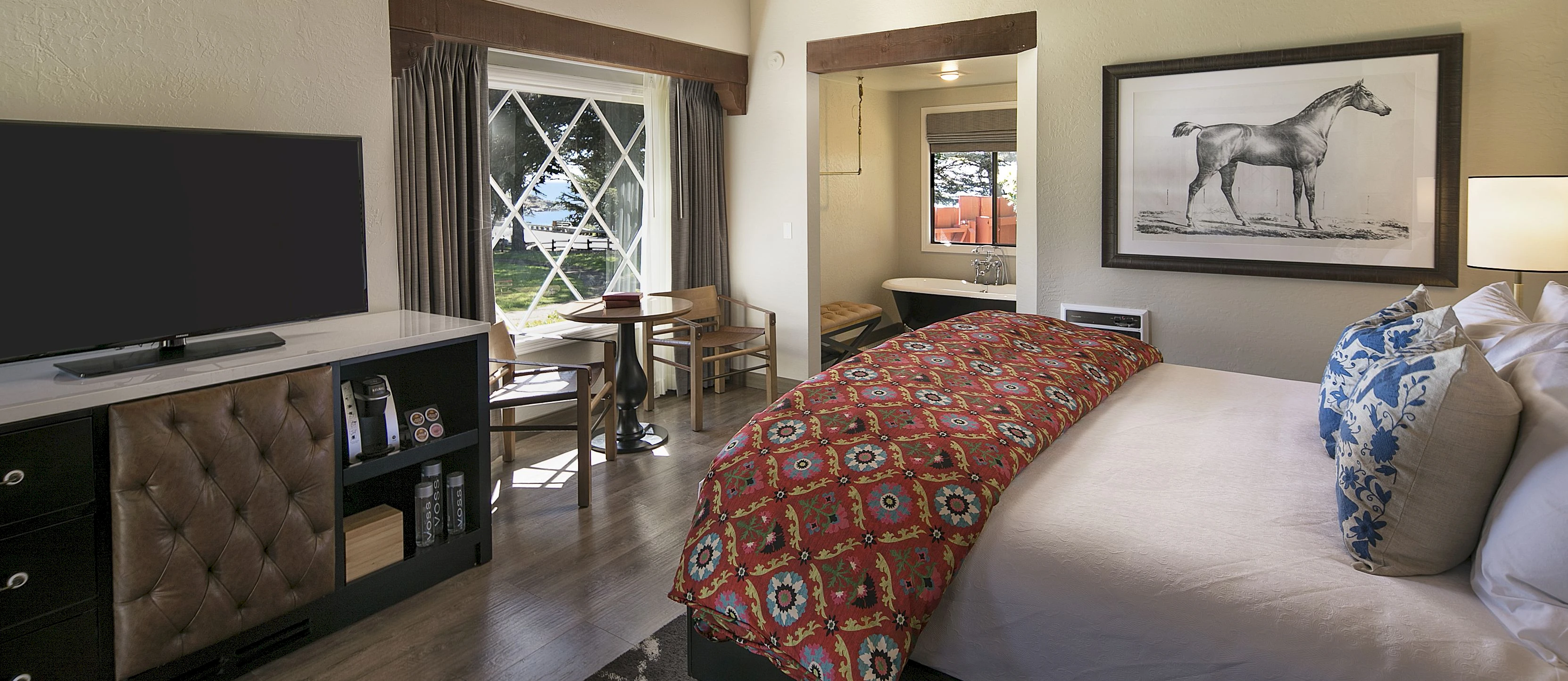 Cozy hotel room with a large bed, patterned bedding, wood ceiling, desk area, TV, and a framed horse artwork on the wall.