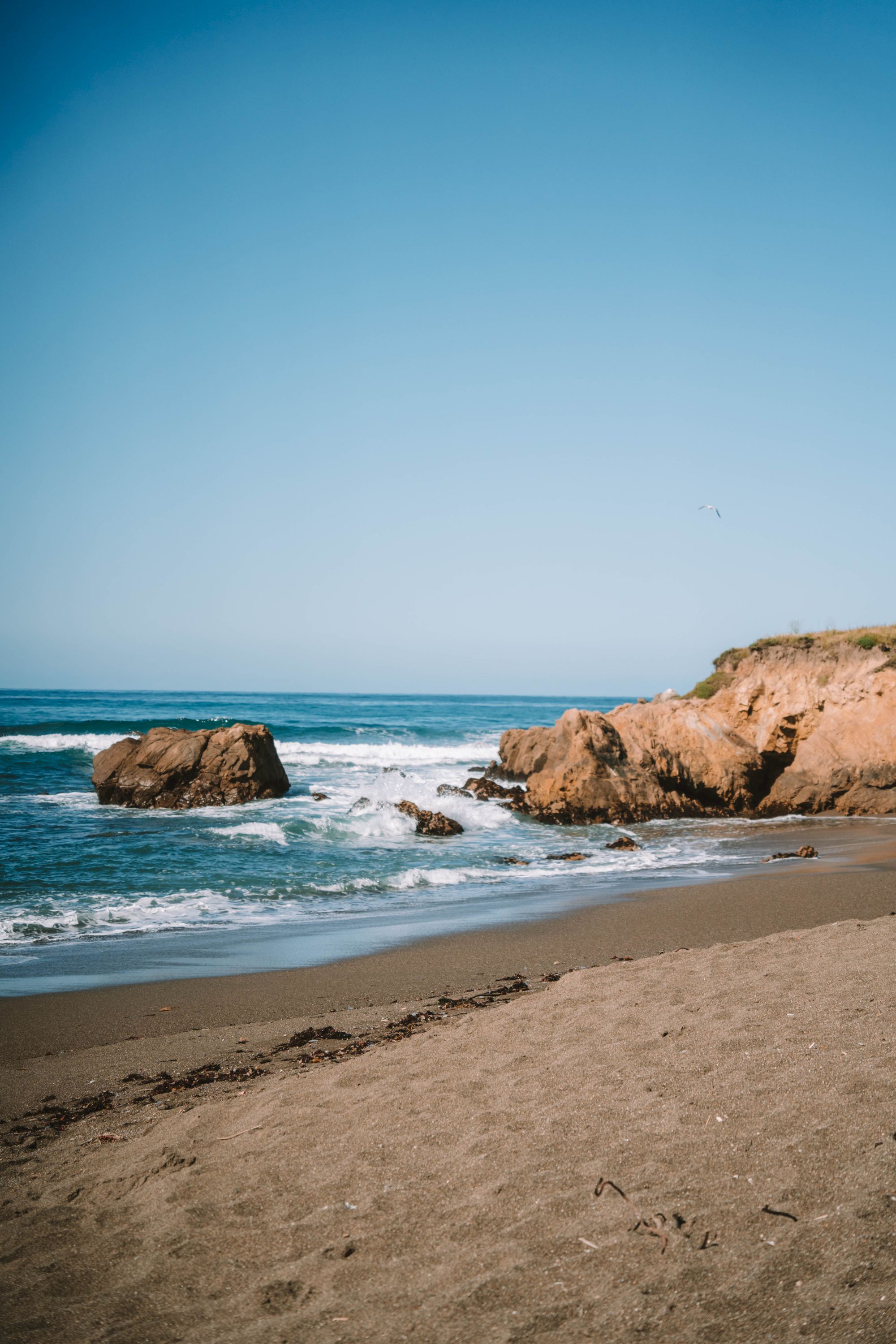 A quiet beach scene with gentle waves, rocky shoreline, and clear blue sky; footprints fade in the warm sand as the ocean lulls the coast to wake.