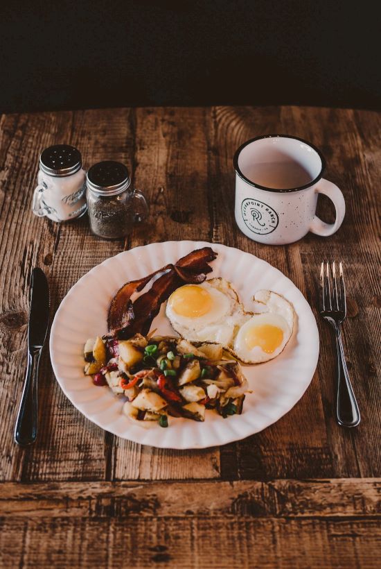 A hearty breakfast with two sunny-side eggs, bacon, saut&eacute;ed vegetables, and potatoes on a white plate, beside a mug and condiments on a wooden table.