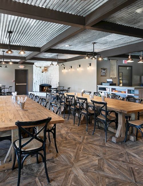 A modern, airy caf&eacute; with long wooden communal tables, metal chairs, industrial ceiling, warm lighting, and a counter with a fridge and display case in the back.