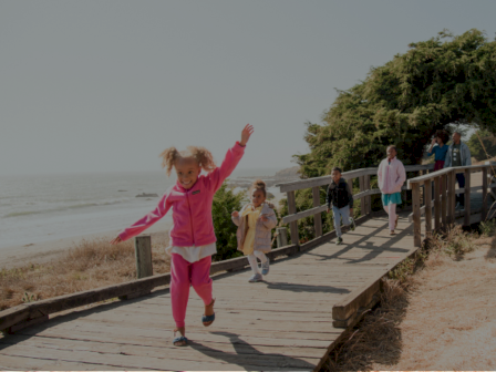 A joyful girl in a pink tracksuit runs excitedly on a wooden boardwalk near the beach, followed by others strolling along the path.