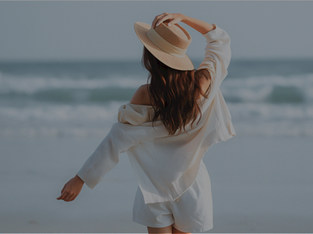 A person in a white outfit and hat stands on a beach, facing the ocean with arms out, enjoying a breezy seaside moment.
