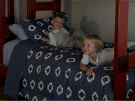 Two kids on a bunk bed, smiling and leaning over the edge of a blue patterned quilt.