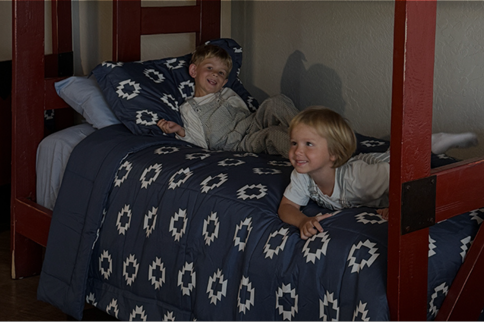 Two young boys lying on a bunk bed with a blue and white comforter and pillows.
