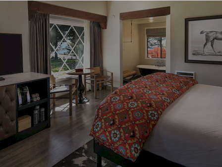 A cozy hotel room with a large bed, patterned red quilt, desk and chair, a TV cabinet, a window with curtains, and a framed horse drawing on the wall.