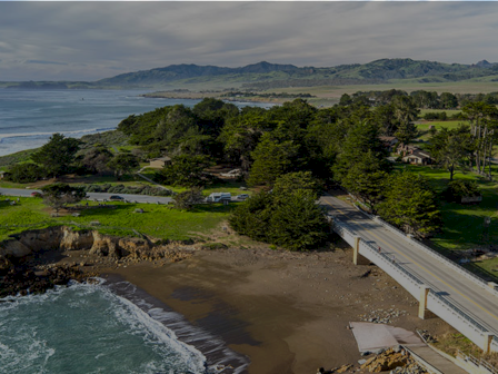 Coastal park with a bridge over a sandy beach, green trees, and rolling hills in the distance by the ocean.