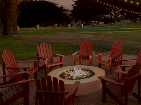 A circle of red Adirondack chairs surrounds a lit fire pit in a park at dusk, with string lights overhead.