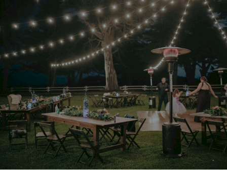 Outdoor nighttime party scene with string lights, tables, chairs, a dance area, and a few guests mingling near a heater, under a tree.