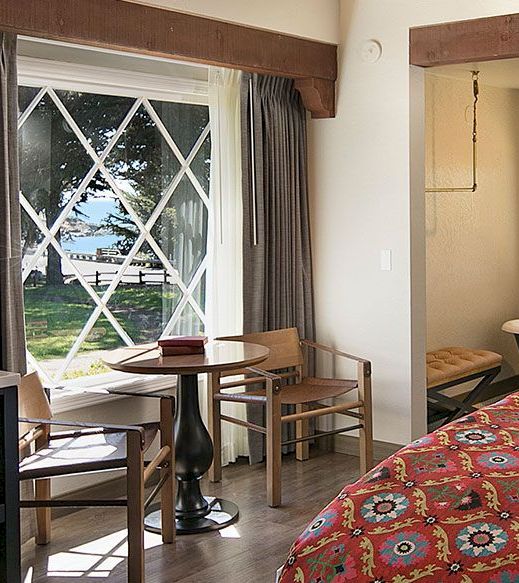A guest room with a large window, a small table with chairs, and a bed with a floral bedspread.