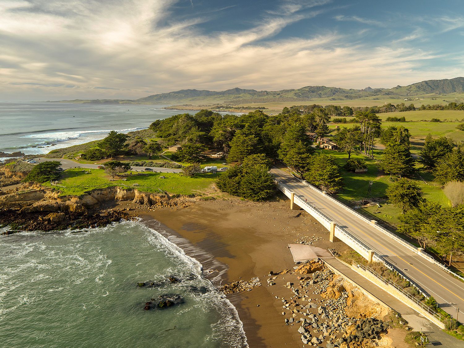 A coastal landscape with a curving road by the beach, waves crashing on rocky shore, green fields, trees, and distant mountains under clouds.