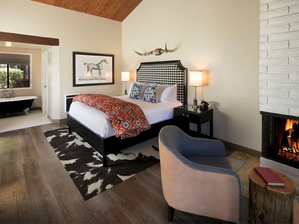 A stylish bedroom with a large bed, plaid headboard, orange throw, nightstands, and a fireplace, plus a seating area and a framed animal art piece.