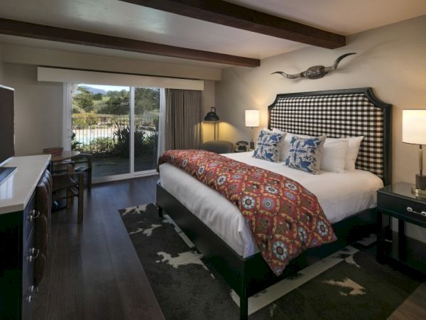 A cozy hotel room with a large bed, checkered headboard, patterned orange throw, two nightstands, lamps, and a sliding door to a patio. ending.
