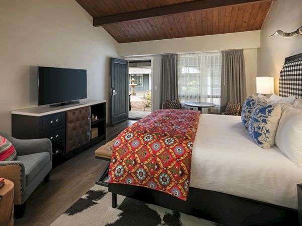 A cozy hotel room with a king bed, colorful patterned throw, sitting area, flat-screen TV, and a balcony view.