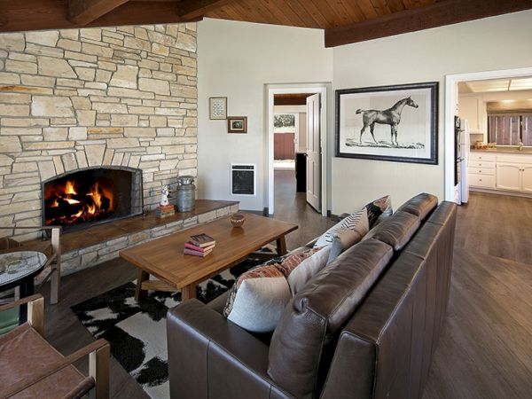 Cozy living room with a stone fireplace, brown leather sofa, wooden coffee table, and a framed horse artwork; warm wood floors and open kitchen in back.