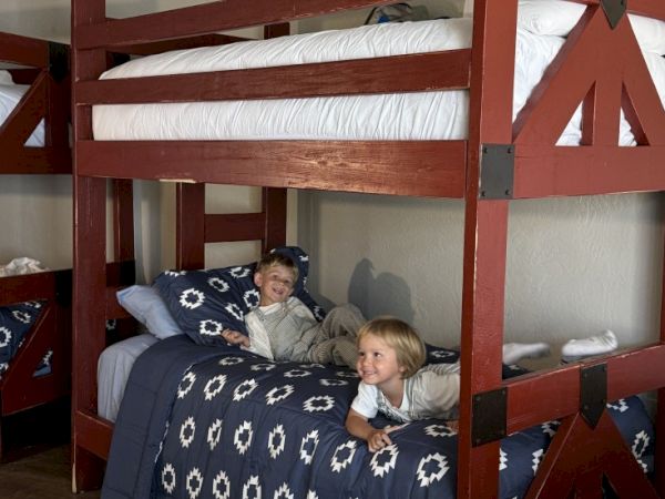 Two kids sit on the lower bunk of a wooden bunk bed in a cozy room, smiling at the camera.