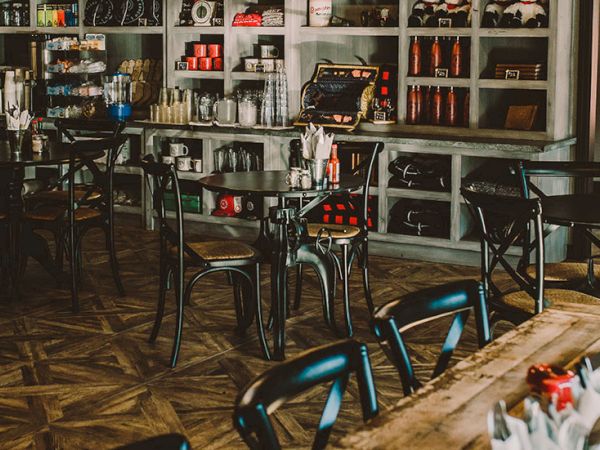 Cozy caf&eacute; with wooden chevron floor, metal chairs, and a long wooden table in the foreground; shelves and drinks behind the counter, warm lighting.