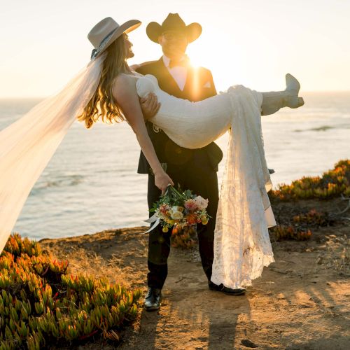 A couple dressed in wedding attire by the sea; the groom lifts the bride as the sun sets, she holds a bouquet and a flowing veil trails behind.
