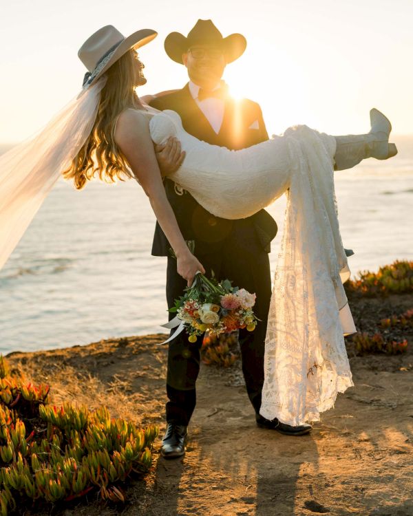 A couple dressed in wedding attire by the sea; the groom lifts the bride as the sun sets, she holds a bouquet and a flowing veil trails behind.