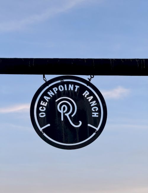 A round hanging sign reads &ldquo;Oceanpoint Ranch&rdquo; with a stylized &ldquo;OR&rdquo; logo, mounted under a beam against a blue sky.