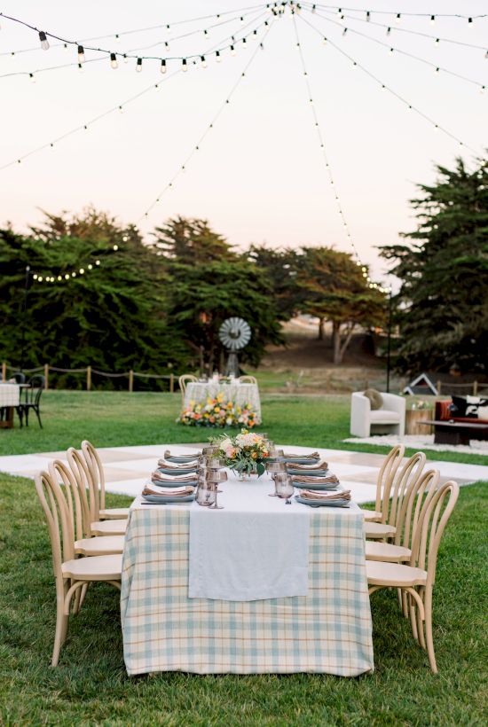 A garden dinner setup with a long table, chairs, string lights overhead, and a decorative centerpiece outdoors.