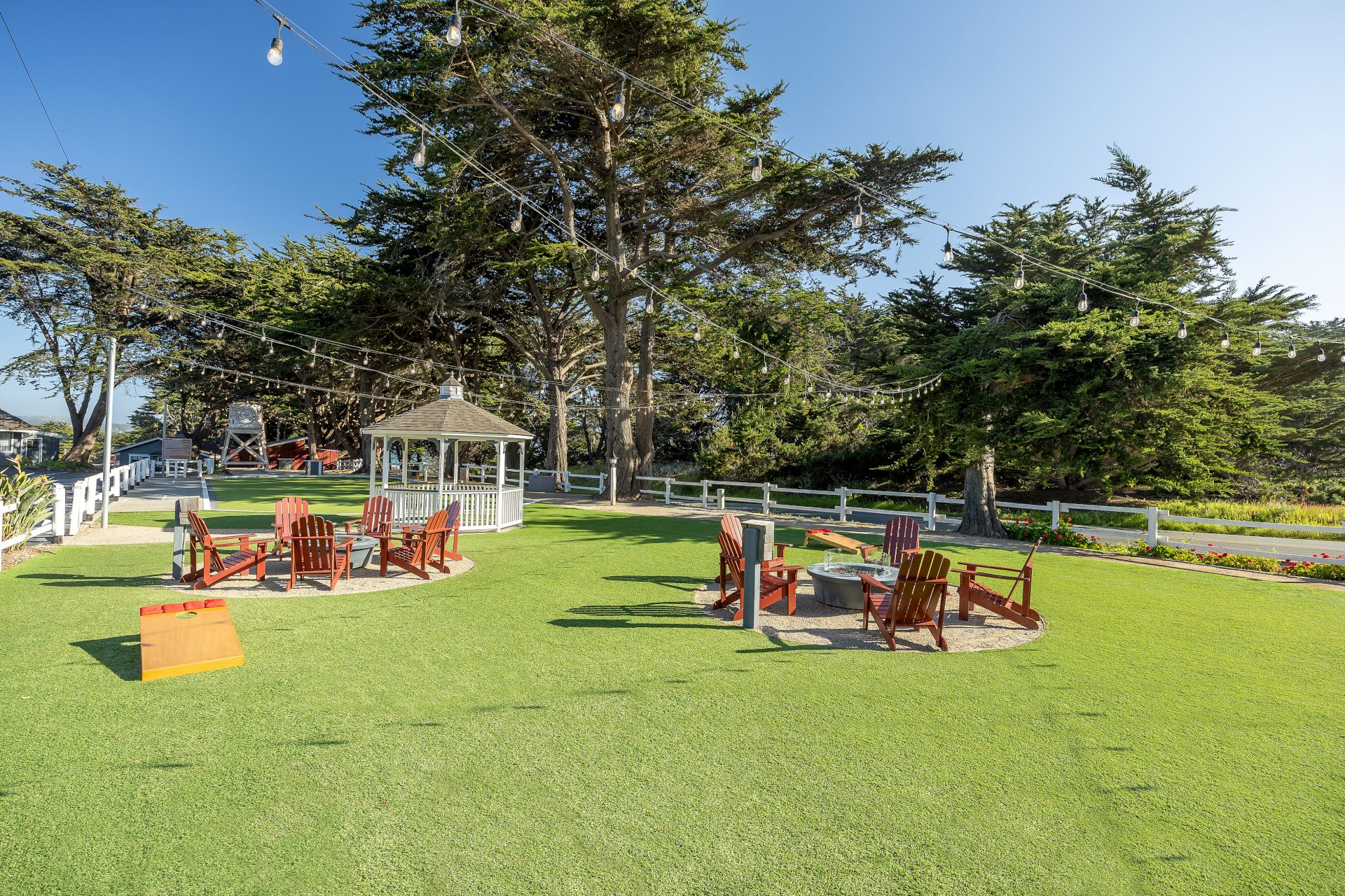 People relaxing on a grassy lawn with Adirondack chairs, a small gazebo, string lights, and tall trees in the background, sunny day.