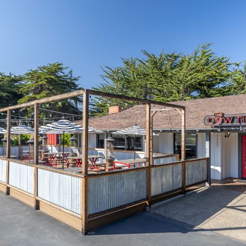 A small outdoor-patio dining area attached to a rustic building with a red door, wooden rails, umbrellas, and blue sky.