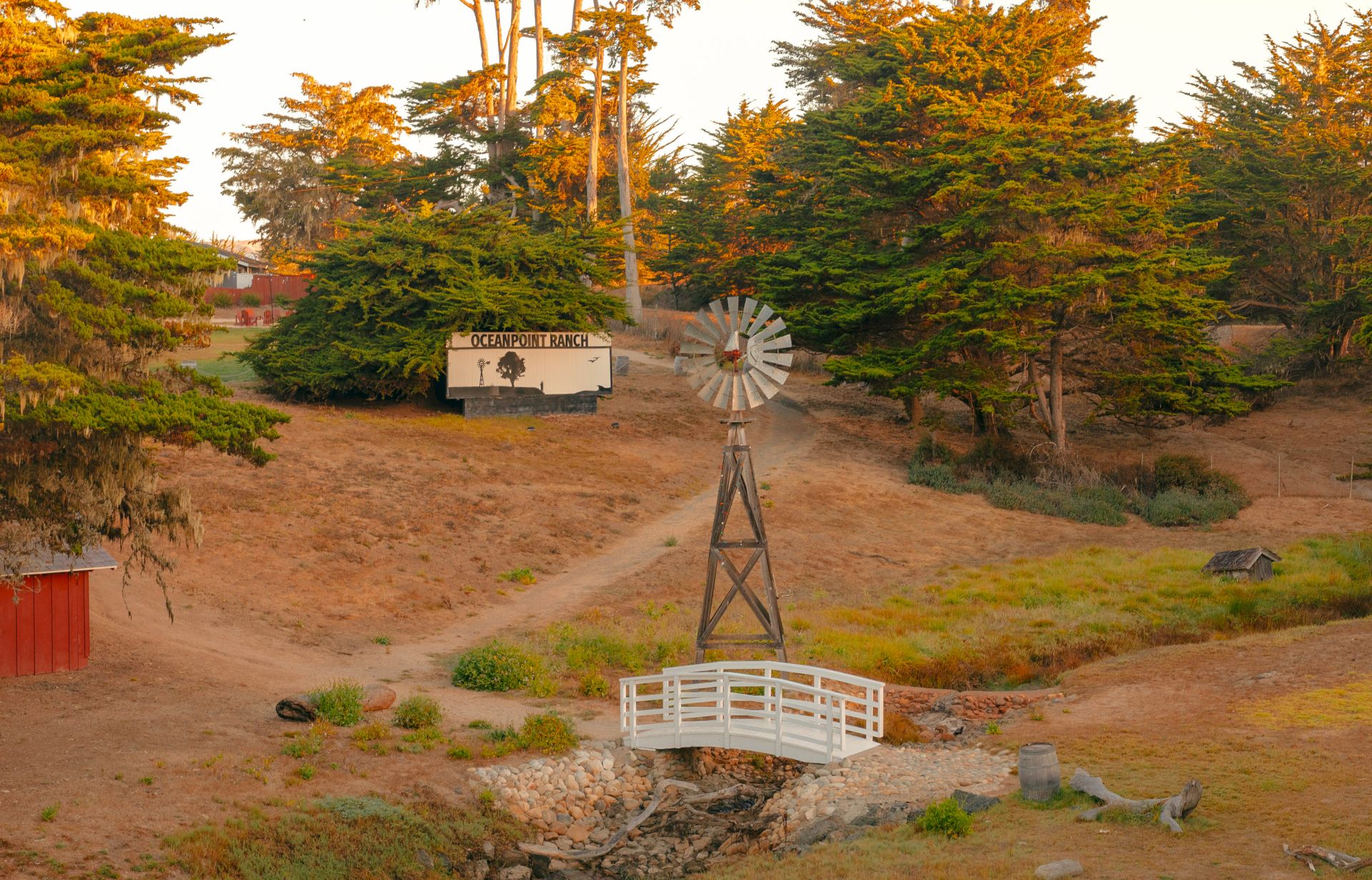 A rural scene with a windmill, small white bridge over a dry creek, dirt paths, pine trees, and a wooden shed in the background.