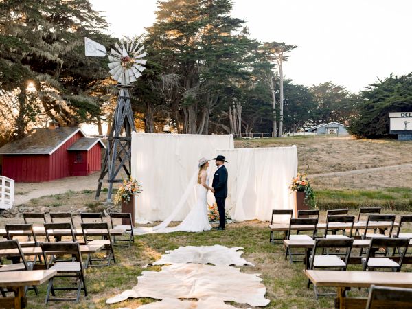 A couple ties the knot at an outdoor rustic wedding setup with a white backdrop, windmill, wooden chairs, and a flowered aisle.
