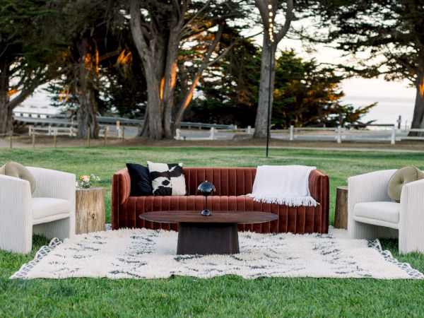 A stylish outdoor seating arrangement on a lawn: a brown sofa with white throw, two white armchairs, a wooden coffee table on a patterned rug, with trees and a fence in the background.