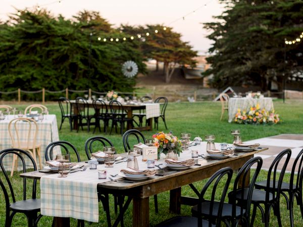 A rustic outdoor reception setup with long wooden tables, mismatched chairs, checkered tablecloths, and string lights in a garden setting, ready for guests.