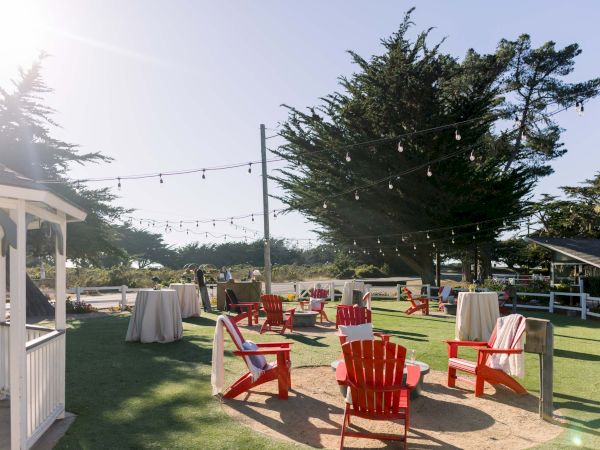 Outdoor party setup with string lights, red adirondack chairs, high-top tables, and a shaded lawn under a large tree.