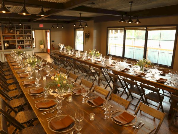 A long wooden banquet table setup with plates, glassware, and napkins, set for a crowd in a warmly lit room with large windows.