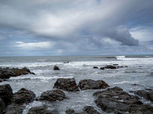 A rocky shoreline with dark jagged stones along the water&rsquo;s edge, waves crashing softly under a cloudy, overcast sky.