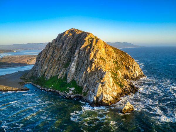 A rocky sea cliff island rising from the ocean, with sunlit, jagged rock face, blue waters, and a sandy beach at its base, surrounded by waves.