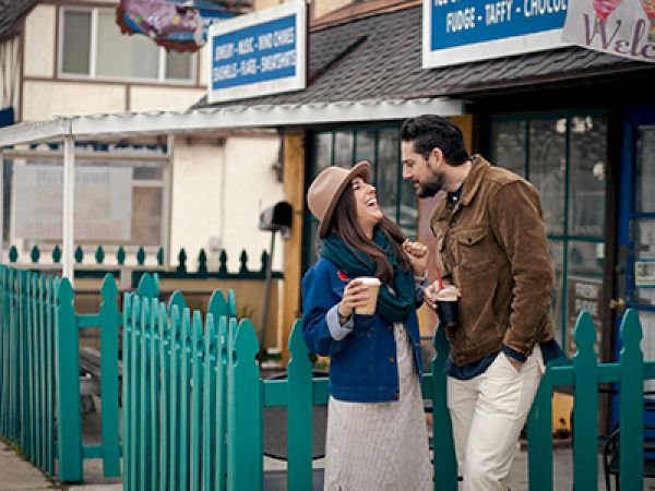 Two people stand outside a cafe, smiling at each other, holding cups and sharing a warm conversation near a teal fence.