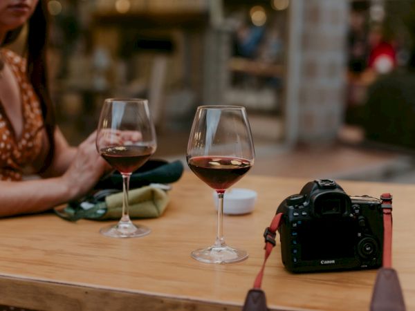 Two wine glasses and a camera on a wooden table, with a person in the background, enjoying a relaxed outdoor setting.