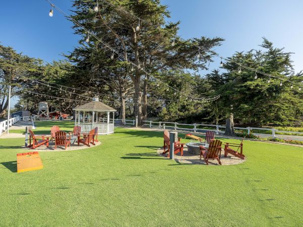A sunny outdoor lawn with string lights, wooden chairs in small circles, a white gazebo, trees in the background, and a calm coastal vibe.