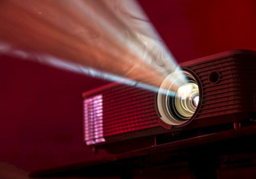 A red projector beams its bright light onto a screen, casting a focused circle as it whirs in a dark, warm-toned room.