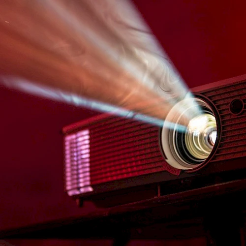 A red projector beams its bright light onto a screen, casting a focused circle as it whirs in a dark, warm-toned room.