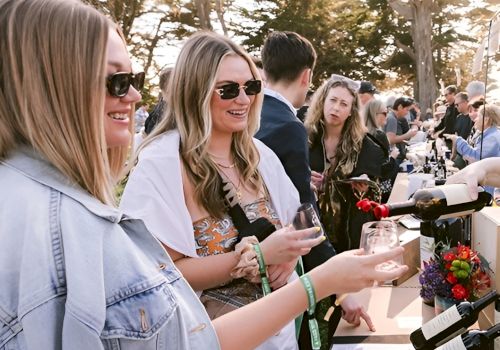 Three friends at a sunny outdoor event, pouring drinks at a bar counter with others chatting nearby and cameras around.