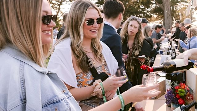 Three friends at a sunny outdoor event, pouring drinks at a bar counter with others chatting nearby and cameras around.