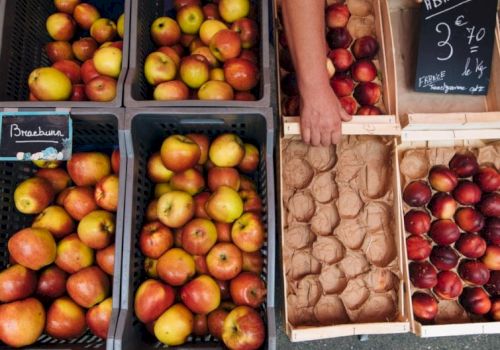 A street market scene with crates of apples and pears on the left, and eggs and peaches in paper trays on the right.