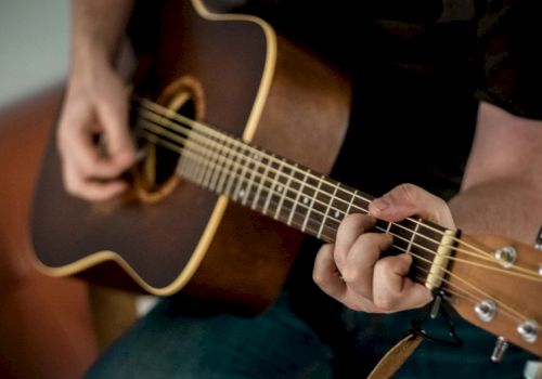 A person is playing a brown acoustic guitar, fingers strumming near the soundhole, focusing on the fretboard as they pick a chord.