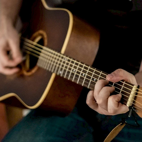 A person is playing a brown acoustic guitar, fingers strumming near the soundhole, focusing on the fretboard as they pick a chord.