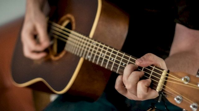 A person is playing a brown acoustic guitar, fingers strumming near the soundhole, focusing on the fretboard as they pick a chord.