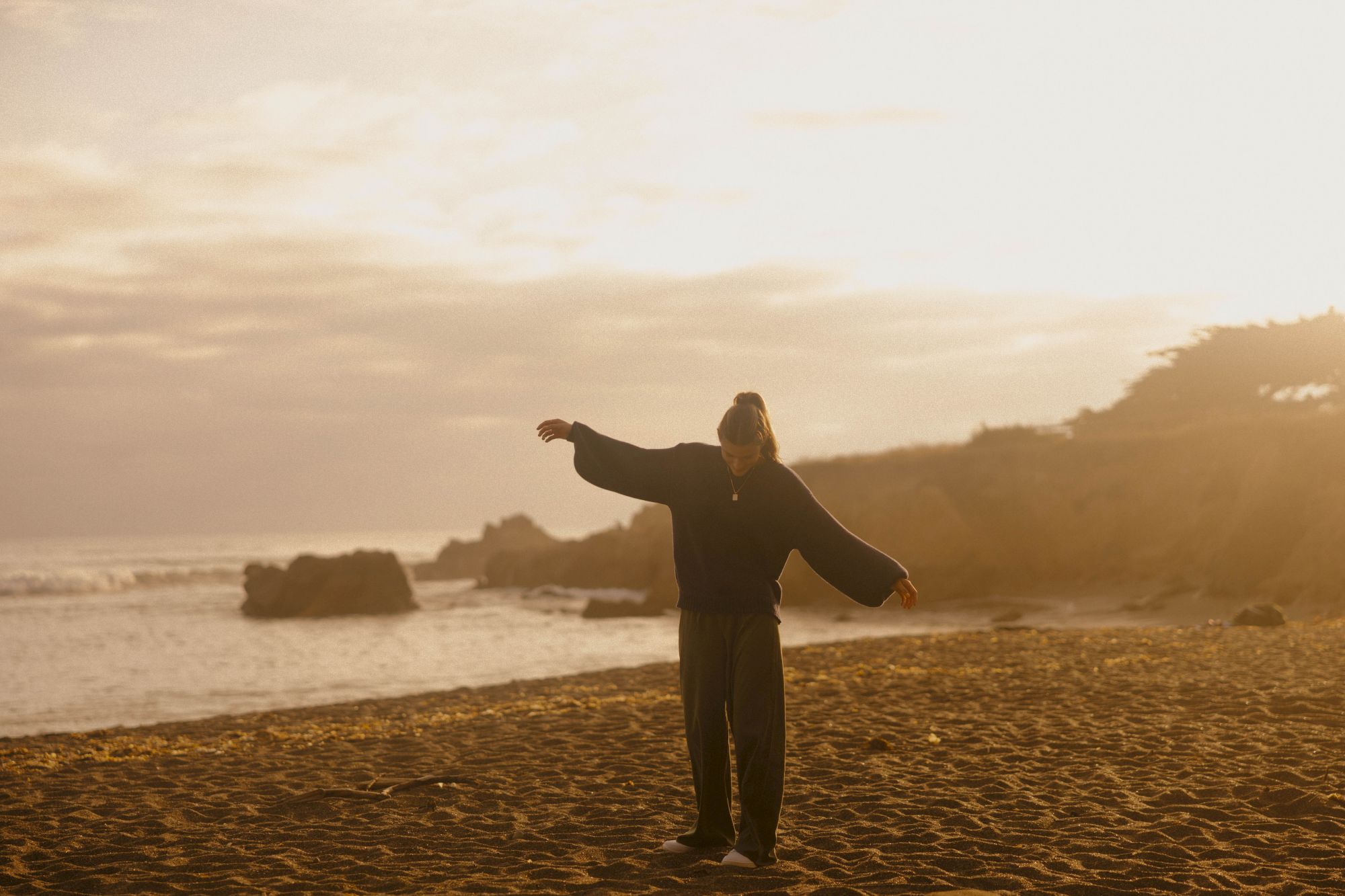A person stands on a sandy beach with arms wide open, facing the waves as the sun sets, painting the scene in warm, golden light.