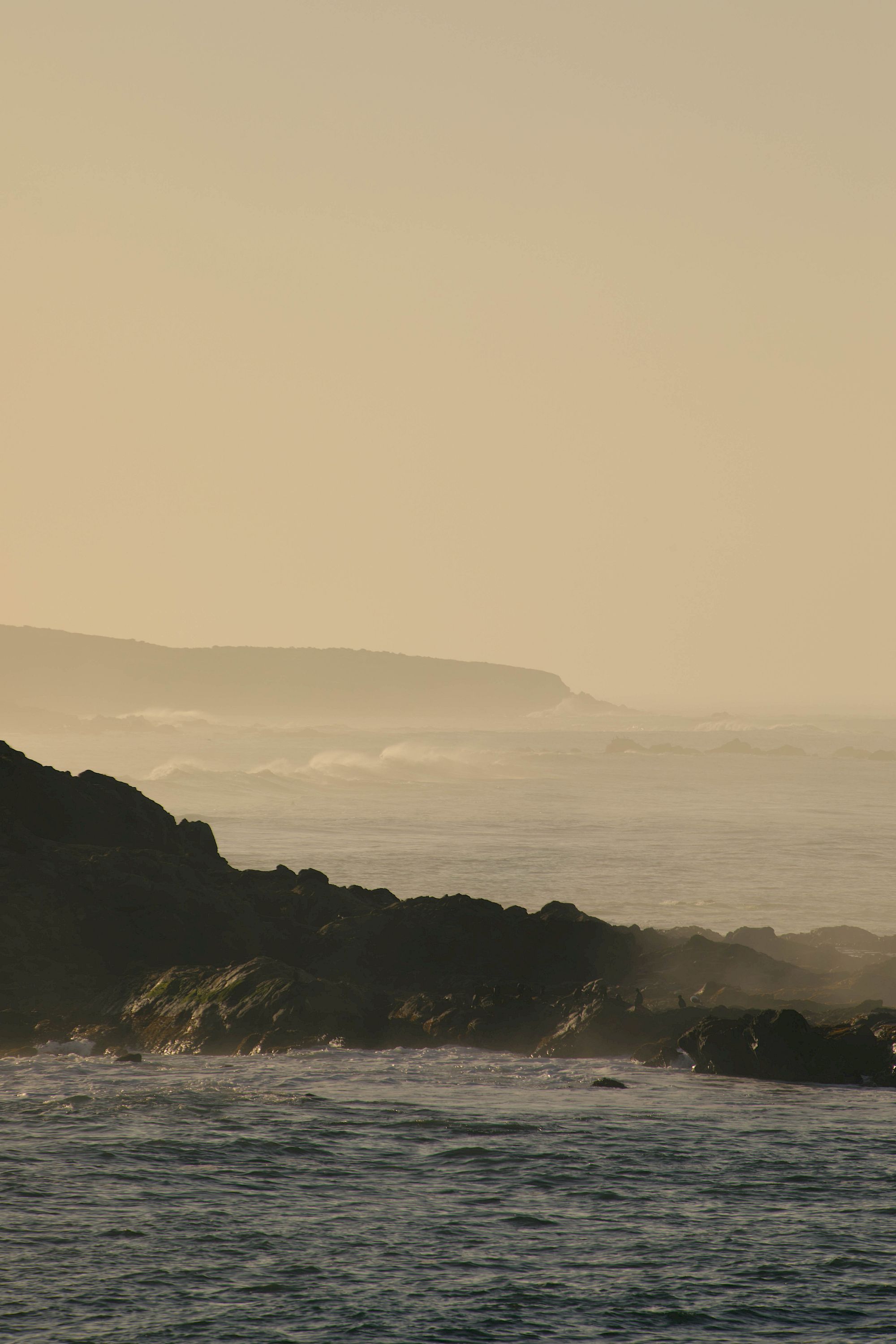 A serene coastal scene at sunset with calm waters, rocky shoreline, and a soft golden sky fading into the horizon.